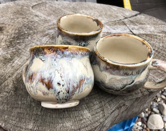 Może przedstawiać: Trzy ceramiczne kubki z białą, brązową i niebieską glazurą. Kubki mają zaokrąglony kształt i uchwyt.