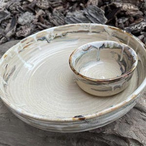 May include: A handmade ceramic serving bowl set. The large bowl is beige with a spiral pattern, and the smaller bowl is nestled inside. Both bowls have a brown and blue rim and a glossy finish.