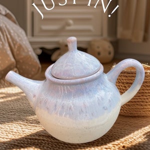 May include: A white ceramic teapot with a light blue glaze and a speckled finish. The teapot has a round body and a curved spout. The teapot is sitting on a woven rug.