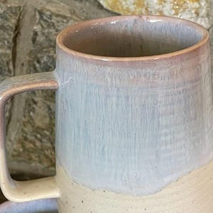 May include: A large, light grey ceramic mug with a light brown handle and a light brown rim. The mug has a light brown glaze on the bottom half and a light grey glaze on the top half.