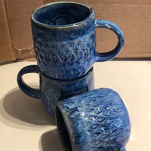 May include: Three blue ceramic mugs with a textured, embossed design. The mugs are stacked on top of each other.