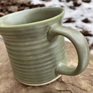 May include: A handmade, sage green ceramic mug with a curved handle. The mug has a ridged texture around the body and a white base. The mug is sitting on a rock, with a blurred background of leaves and snow.