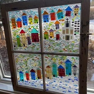 May include: A stained glass window depicting a village scene. The window shows colourful houses with red, blue, green, and yellow roofs. A white church with a cross is also shown. The background is a mix of blue, green, and white glass.