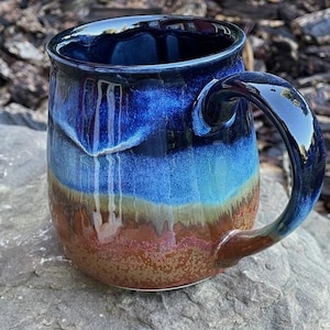 May include: A ceramic mug featuring a reactive glaze with a deep navy blue rim that transitions into a vibrant sky blue middle section, ending in a textured, earthy brown base. The mug has a curved handle and a smooth, glossy finish throughout.