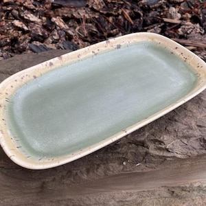 May include: An oval ceramic tray featuring a smooth, matte sage green center basin. The raised rim is finished in a contrasting cream glaze with dark brown speckled accents distributed along the edges, creating a textured appearance.