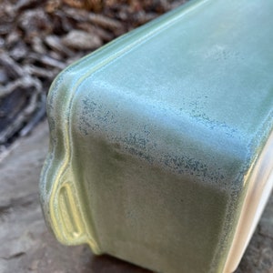 May include: A green ceramic baking dish with a speckled glaze. The dish has a rectangular shape with rounded edges and a slightly raised rim.