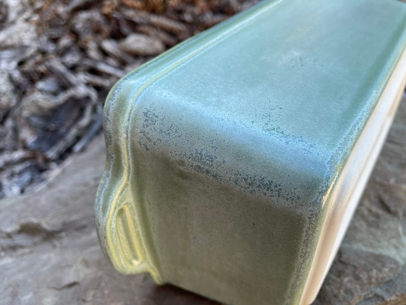 May include: A green ceramic baking dish with a speckled glaze. The dish has a rectangular shape with rounded edges and a slightly raised rim.