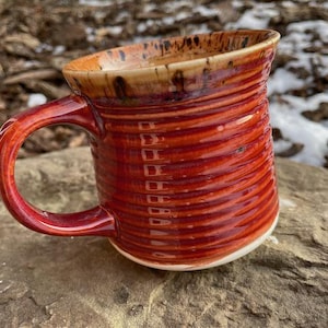 Peut inclure: Une tasse en céramique faite à la main avec un glaçage rouge et brun. La tasse a une base large et arrondie et un bord légèrement évasé. La poignée est fixée sur le côté de la tasse et est également glacée en rouge.