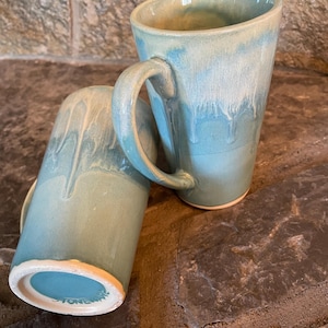Peut inclure: Deux tasses en céramique avec un glaçage bleu clair et des stries blanches. Les tasses sont faites à la main et ont un aspect rustique. Le fond d'une tasse porte l'inscription "Stoneware".