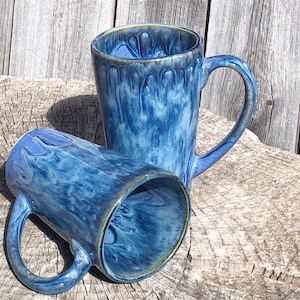 May include: Two blue ceramic mugs with a marbled glaze pattern. The mugs have a tall, cylindrical shape and a handle on one side.