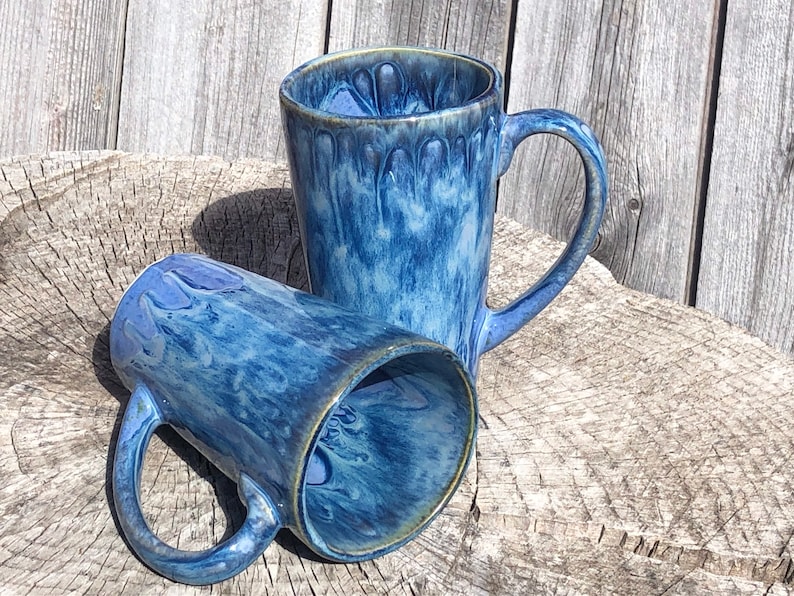 May include: Two blue ceramic mugs with a marbled glaze pattern. The mugs have a tall, cylindrical shape and a handle on one side.