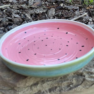 May include: A round ceramic bowl with a smooth, glossy pink interior featuring small, scattered black dots and concentric circular ridges. The exterior of the bowl has a gradient finish transitioning from a light green to a soft blue hue.