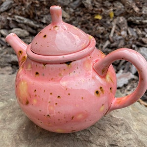 May include: A pink ceramic teapot with a speckled glaze and a lid. The teapot has a round body and a curved handle.