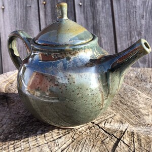 May include: A handcrafted ceramic teapot with a lid, handle, and spout. The teapot has a glossy finish with shades of green, blue, and brown. It is sitting on a wooden surface, showcasing its unique, artisanal design.