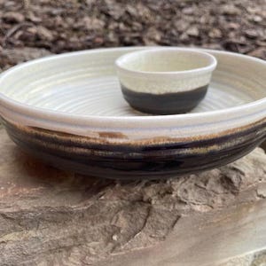 May include: A handmade ceramic chip and dip serving set. The large bowl has a cream-coloured interior and a dark brown exterior. A smaller dip bowl sits in the centre. The set is on a natural stone surface.