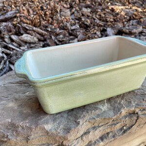 May include: A light green ceramic loaf pan with a white interior. The pan has two handles on the sides.