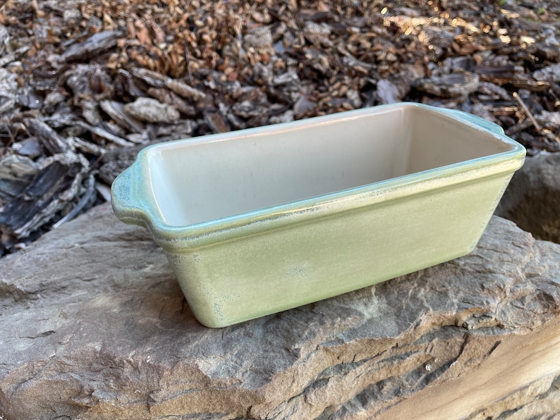 May include: A light green ceramic loaf pan with a white interior. The pan has two handles on the sides.