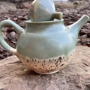 May include: A green ceramic teapot with a speckled glaze and a matching lid. The teapot has a rounded body and a spout on one side. The lid has a small knob on top.