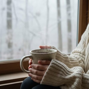 May include: A speckled off-white ceramic mug with a wide handle and a slightly flared rim. The mug has a textured, rustic appearance with small dark flecks distributed across its surface, containing a dark liquid with visible steam rising.