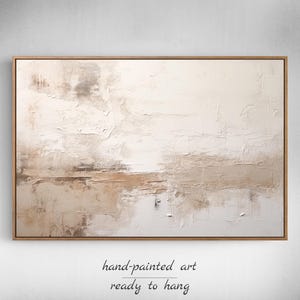 Op de afbeelding: Abstract schilderij in beige en witte tinten. Het schilderij heeft een textuur en is ingelijst in een licht houten lijst. Het schilderij is klaar om opgehangen te worden.