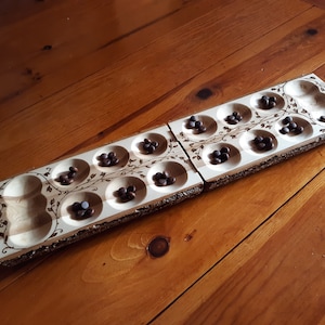 May include: A wooden Mancala board game with two hinged sections. Each section has six circular pits and a larger end pit. The board is decorated with a vine design and filled with brown game pieces. The board is on a wooden surface.