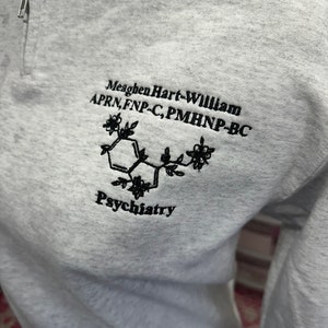 May include: Gray sweatshirt with a black embroidered design. The design includes a stylized chemical structure with flowers and the text "Meaghen Hart-William APRN, FNP-C, PMHNP-BC Psychiatry".