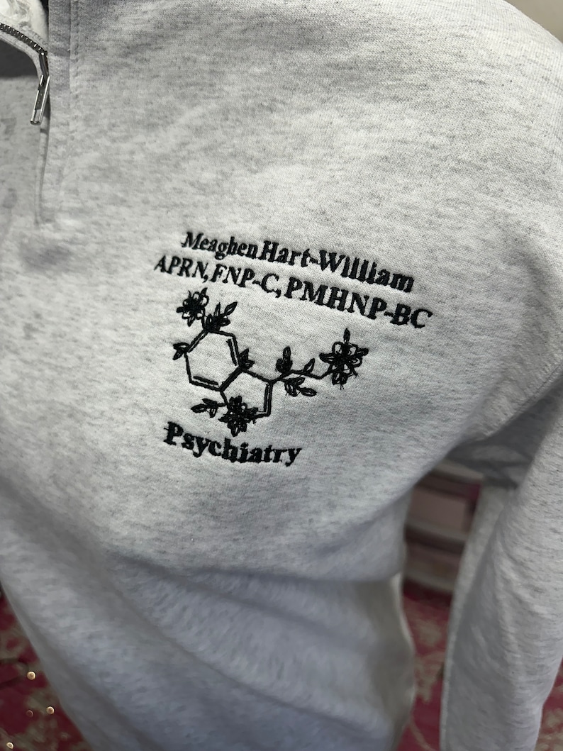 May include: Gray sweatshirt with a black embroidered design. The design includes a stylized chemical structure with flowers and the text "Meaghen Hart-William APRN, FNP-C, PMHNP-BC Psychiatry".