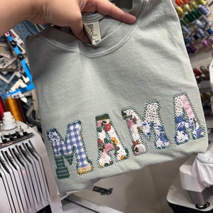 May include: A light green t-shirt with the word "MAMA" spelled out in colorful fabric patches. The patches are sewn together to create a unique and personalized design.