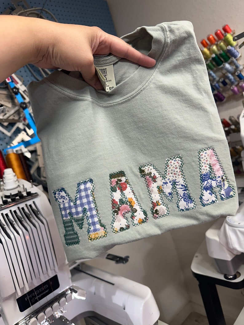 May include: A light green t-shirt with the word "MAMA" spelled out in colorful fabric patches. The patches are sewn together to create a unique and personalized design.