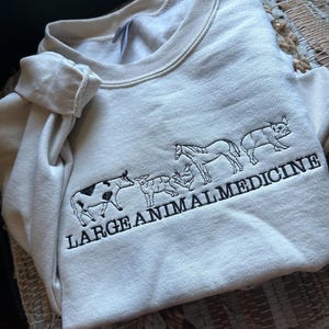 Può includere: Una felpa color crema con un disegno ricamato nero di una mucca, una pecora, un cavallo e un maiale con il testo "LARGE ANIMAL MEDICINE".