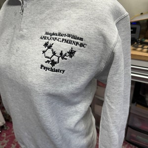 May include: Gray sweatshirt with a black embroidered design. The design includes a chemical structure, the text "Meaghen Hart-William APRN, FNP-C, PMHNP-BC", and the word "Psychiatry".
