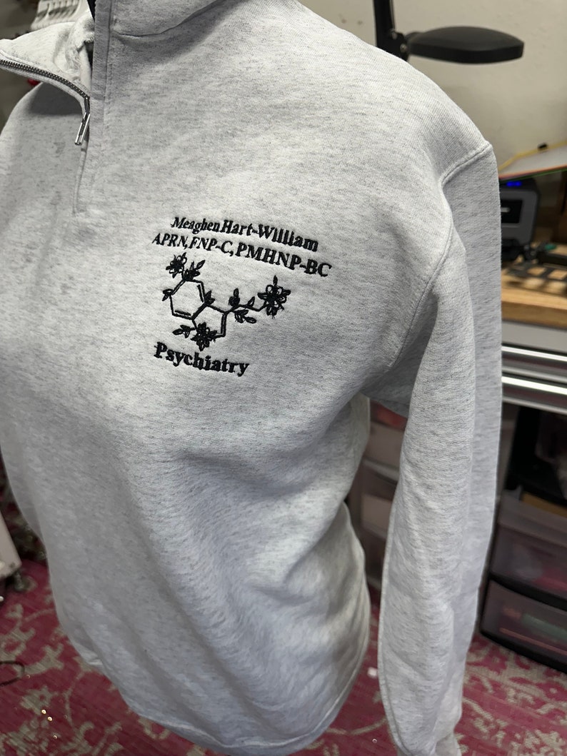 May include: Gray sweatshirt with a black embroidered design. The design includes a chemical structure, the text "Meaghen Hart-William APRN, FNP-C, PMHNP-BC", and the word "Psychiatry".