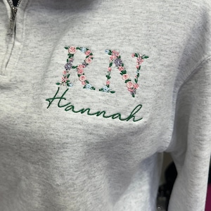 May include: A grey sweatshirt with a zip and a floral embroidered "RN" with the name "Hannah" embroidered below in green.
