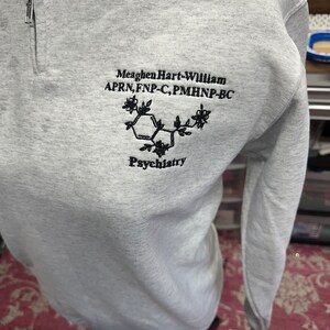 May include: A gray sweatshirt with a black embroidered design. The design includes the text "Meaghen Hart-William APRN, FNP-C, PMHNP-BC" and a stylized chemical structure with the word "Psychiatry" below it.