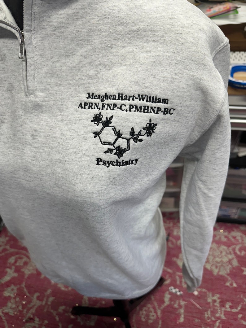 May include: A gray sweatshirt with a black embroidered design. The design includes the text "Meaghen Hart-William APRN, FNP-C, PMHNP-BC" and a stylized chemical structure with the word "Psychiatry" below it.
