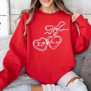 Può includere: Una felpa rossa con una grafica bianca di due cuori collegati da un fiocco. I cuori contengono le parole "XO" e "chia". La felpa è di colore rosso unito.