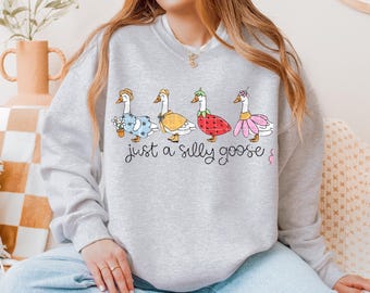 May include: A gray sweatshirt with a graphic of four geese wearing different outfits. The geese are lined up in a row and the text "just a silly goose" is printed below them.