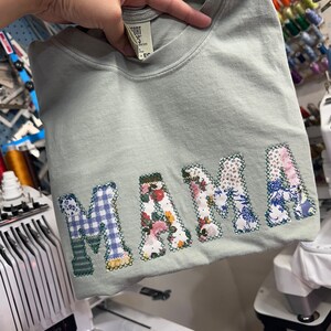 May include: A light green t-shirt with the word "MAMA" spelled out in large, colorful patchwork letters. The letters are made from different fabrics, including floral prints, plaid, and blue and white patterns.