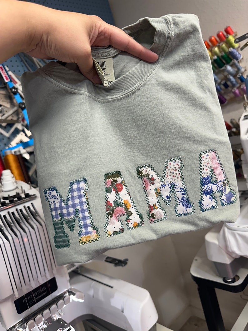 May include: A light green t-shirt with the word "MAMA" spelled out in large, colorful patchwork letters. The letters are made from different fabrics, including floral prints, plaid, and blue and white patterns.