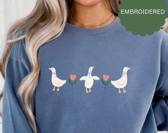 Könnte beinhalten: Ein blaues Sweatshirt mit einem gestickten Design aus drei weißen Enten mit blauen Schleifen, die jeweils vor einer rosa Blume mit grünen Blättern stehen. Der Text "EMBROIDERED" ist in der oberen rechten Ecke des Bildes sichtbar.