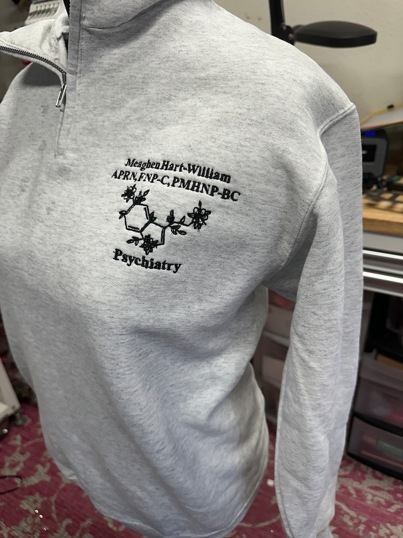 May include: A gray sweatshirt with a black embroidered design. The design includes the text "Meaghen Hart-William APRN, FNP-C, PMHNP-BC" and a stylized chemical structure with the word "Psychiatry" below it.