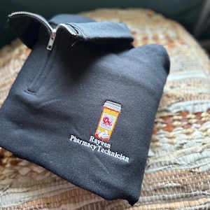 Puede incluir: Sudadera negra con cremallera con una imagen bordada de un frasco de pastillas y el texto "Rayven Pharmacy Technician".