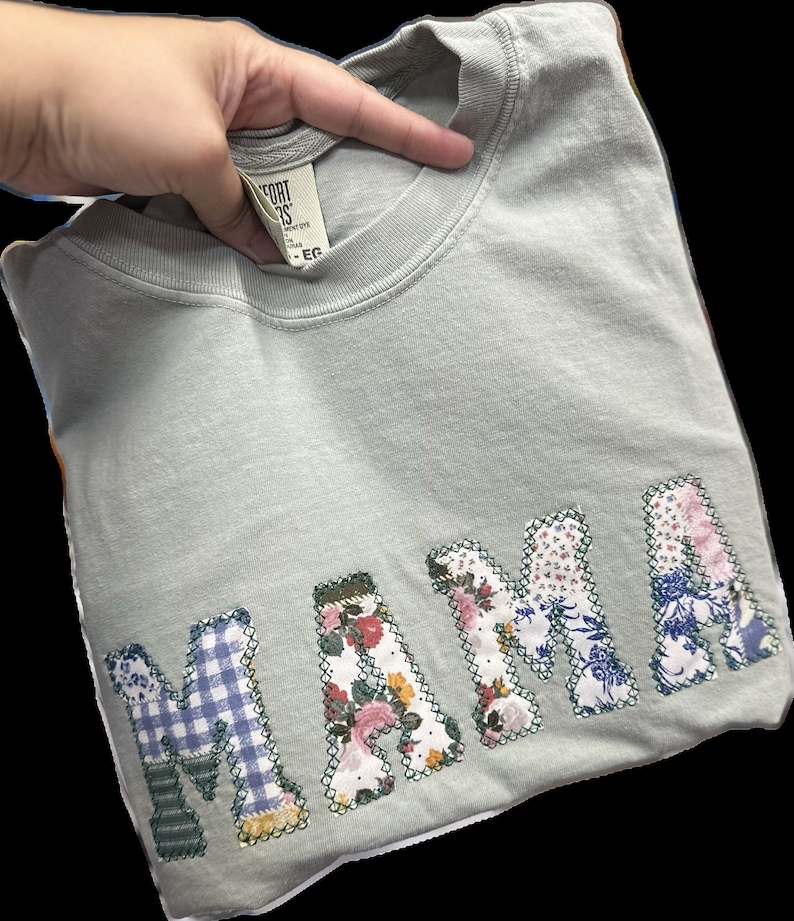 May include: A light blue t-shirt with the word "MAMA" spelled out in large, colorful patchwork letters. Each letter is made from different fabrics with floral and plaid patterns.