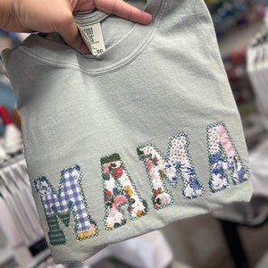May include: A light green t-shirt with the word "MAMA" spelled out in large, colorful patchwork letters. Each letter is made from different fabrics with floral and plaid patterns.