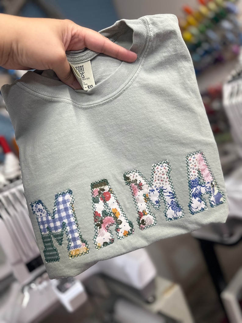 May include: A light green t-shirt with the word "MAMA" spelled out in large, colorful patchwork letters. Each letter is made from different fabrics with floral and plaid patterns.