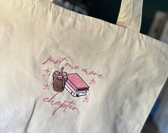 Pode incluir: Saco tote creme com design bordado. O design apresenta uma bebida de café, uma pilha de livros rosa e o texto "just one more chapter" em escrita rosa. O saco tem duas alças creme.