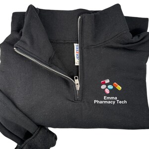 Puede incluir: Sudadera negra con cremallera de cuarto de cremallera, cremallera plateada y diseño de pastillas bordadas. El texto "Emma Pharmacy Tech" está debajo. Fabricado por Jerzees.