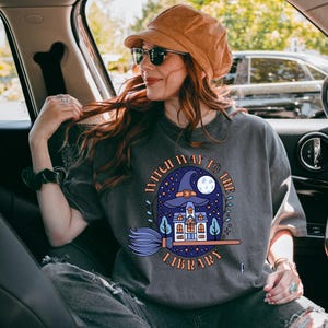 Puede incluir: Una camiseta gris con un gráfico de una bruja volando en una escoba hacia una casa con el texto "Witch Way to the Library".
