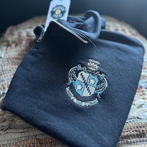 Może przedstawiać: Czarna bluza zapinana na zamek błyskawiczny z haftowanym niebiesko-białym herbem przedstawiającym koronę, tarczę i napis "1820 1920 1980".