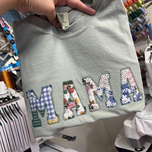 May include: A light green t-shirt with the word "MAMA" spelled out in large, colorful letters. The letters are made of different fabrics, including floral print, plaid, and solid colors.